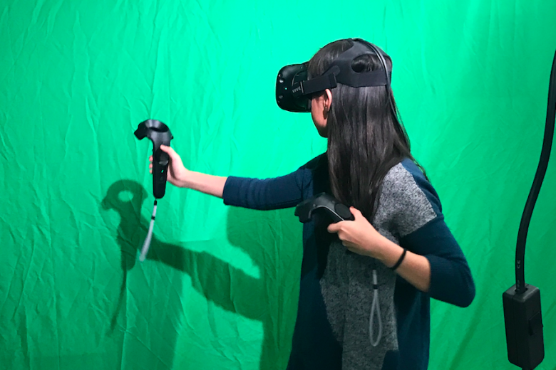 Student wears VR goggles and holds video game controllers in front of green screen