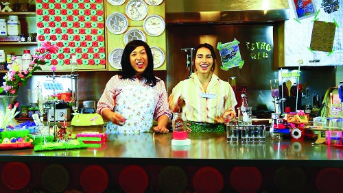 Still from "Housewives Making Drugs" performance piece, two performers stand in neon-colored kitchen with test tube set