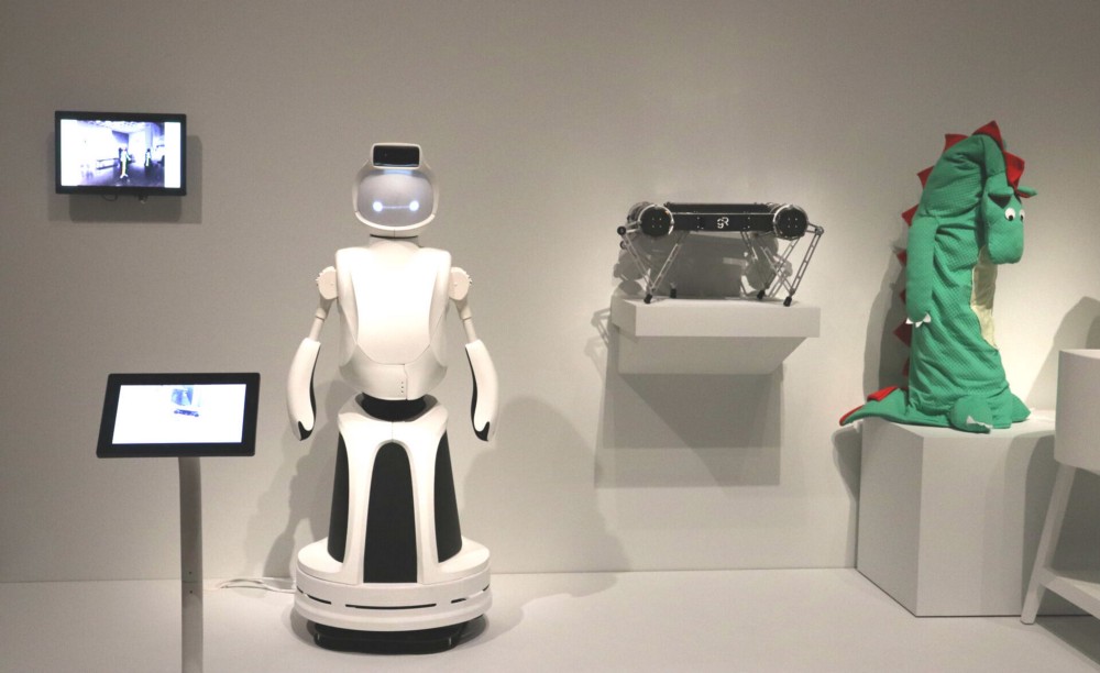 Quori robot, white humanoid robot with digital face, featured in museum exhibit next to Ghost Minitaur, a four legged walking robot, and "Raising Robot Natives", a robot arm covered in green cartoon dragon costume