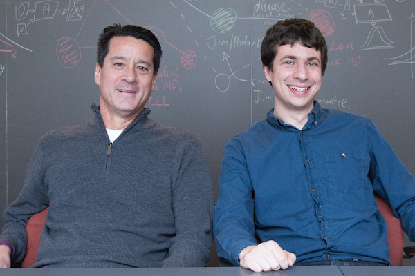 Michael Kearns and Aaron Roth sit in front of a blackboard filled with network diagrams.