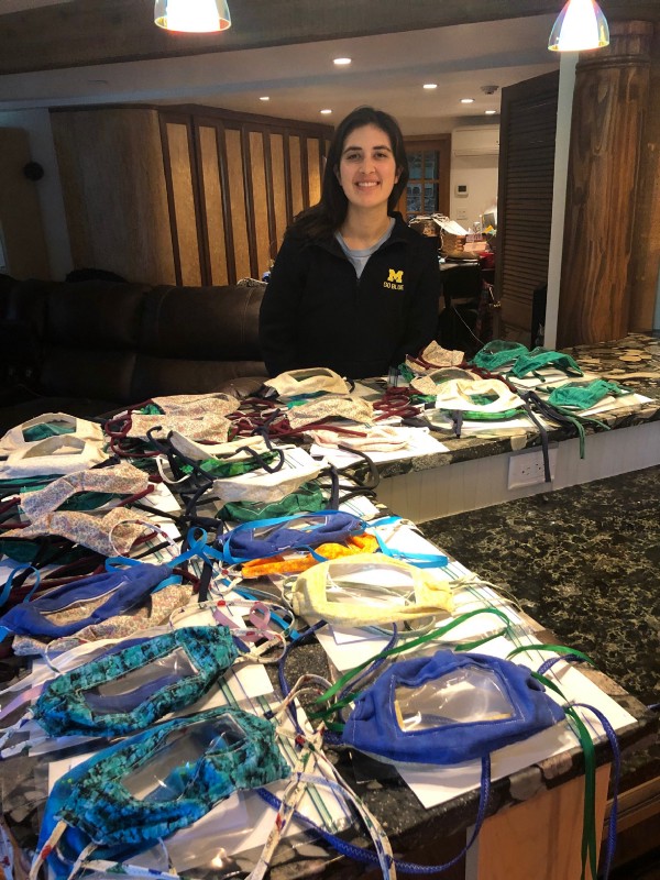 Kate stands in front of many face masks displayed on kitchen counter. Face masks are made of fabric and feature a clear plastic panel in front of mask.