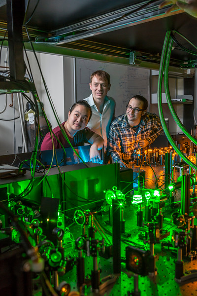 Tzu-Yung Huang, Lee Bassett and David Hopper look on at table where lasers bounce between prisms, lenses and mirrors.