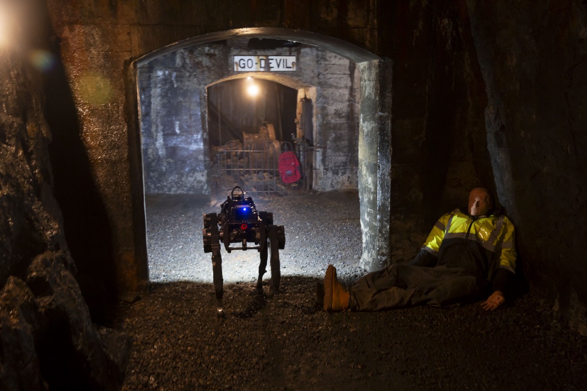 In a dimly lit tunnel, a dog-like legged robot walks past a mannequin posing as a mine-worker in need of rescue.
