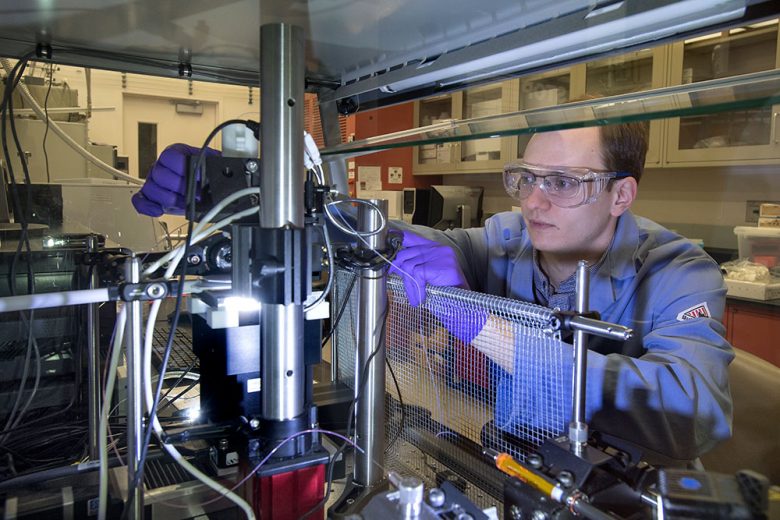 A researcher in a lab coat, goggles and nitrile gloves inspects a piece of complicated machinery.