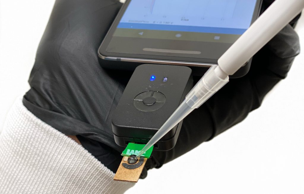 A gloved hand holds a device connected to a cellphone as a pipette deposits a sample onto it.