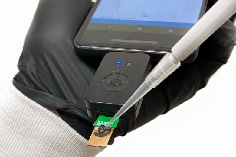 A gloved hand holds a device connected to a cellphone as a pipette deposits a sample onto it.