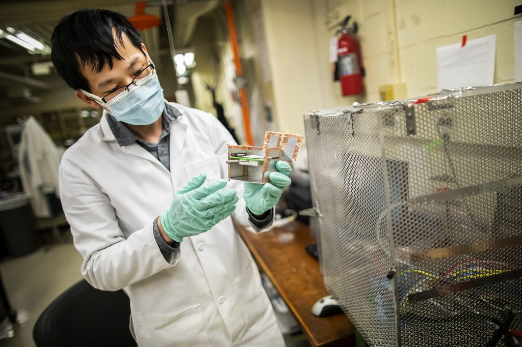 A masked student in a lab coat and gloves examines an electronic device. 