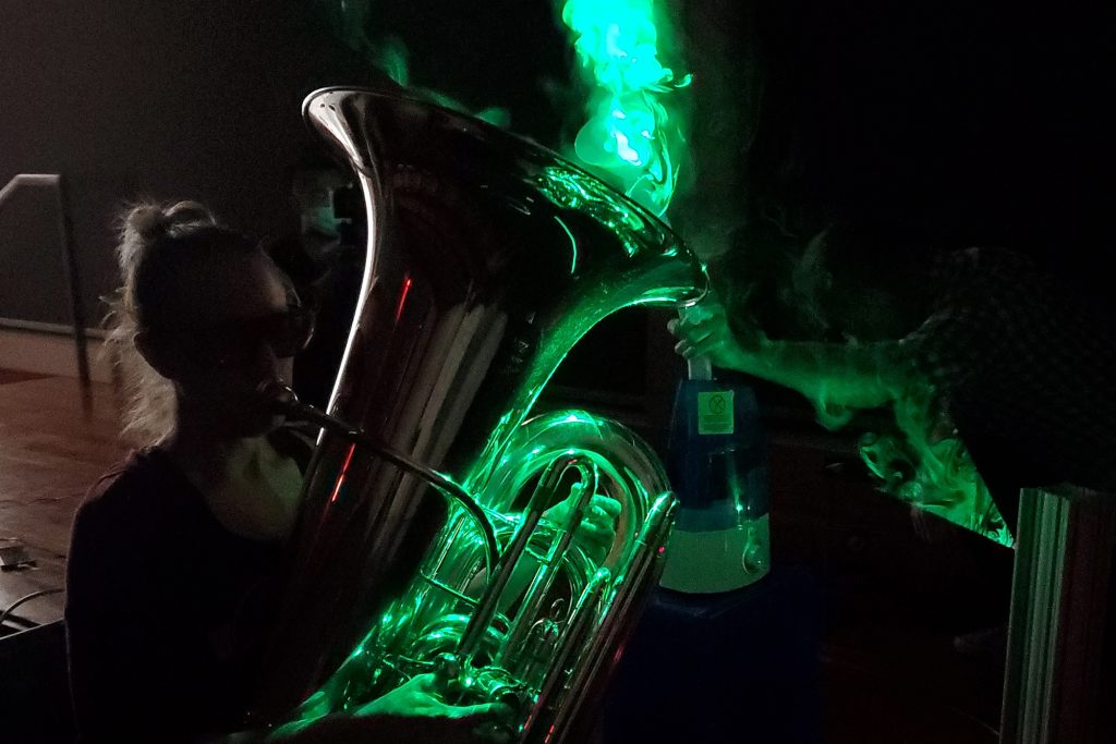 A tuba player bathed in neon green light, as the droplets coming out of the instrument are illuminated by a laser.