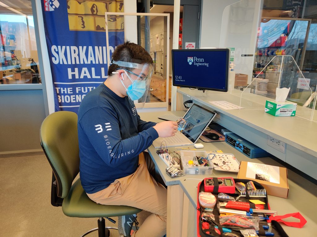 A student in a face mask and shield works at a lab bench covered with electronic equipment.