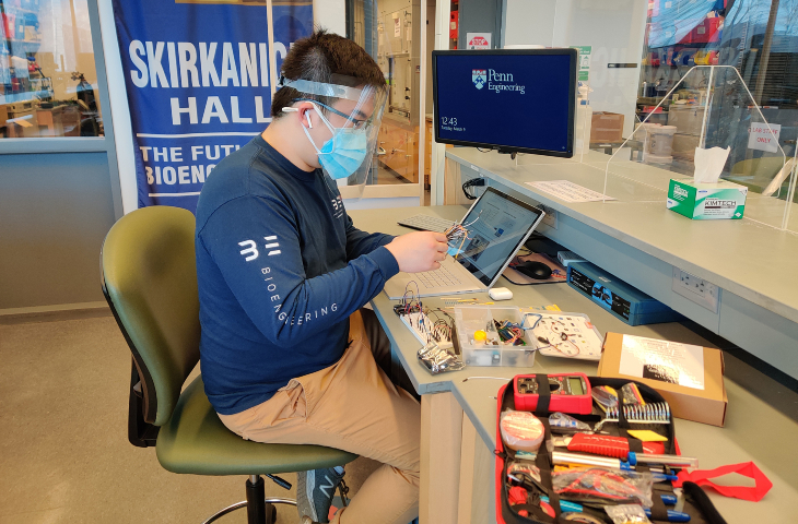 A student in a face mask and shield works at a lab bench covered with electronic equipment.