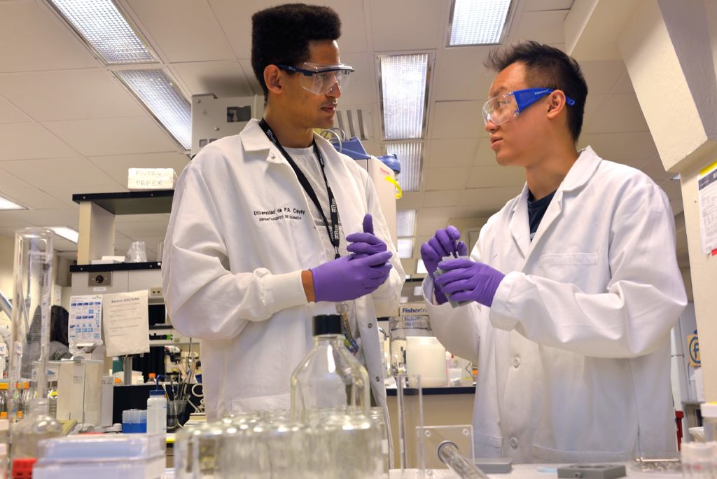 Two researchers in lab coats and purple gloves speak to one another in lab.