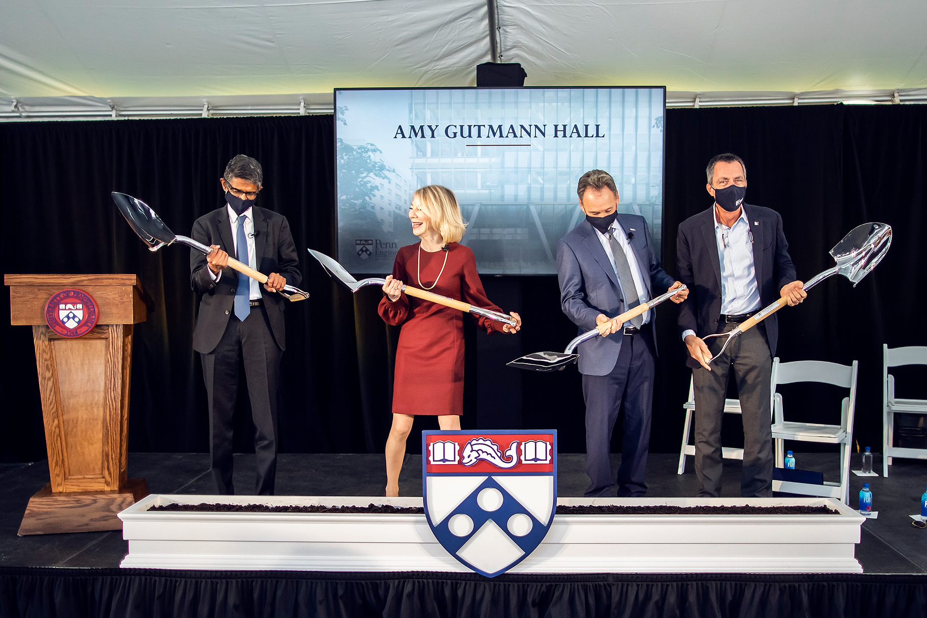 vijay kumar, amy gutmann, Harlan Stone, and Rob Stavis with shovels digging into a trough of dirt on a stage with amy gutmann hall on a screen behind them