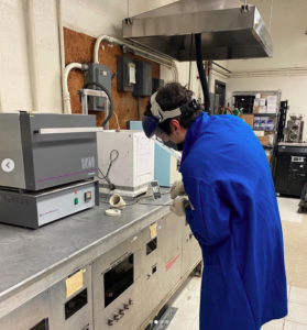 A student in a blue lab coat and face shield pours molten metal into a mold with tongs.