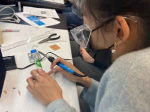 A woman in safety goggles holding a soldering iron and a small plastic tube.