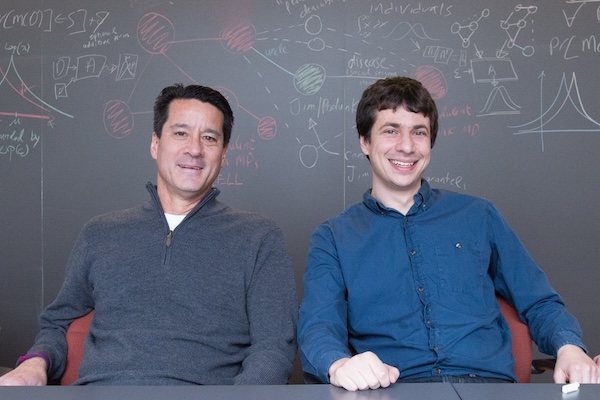 Michael Kearns and Aaron Roth sit side-by-side in front of a blackboard covered in math- and computer science-related notes.