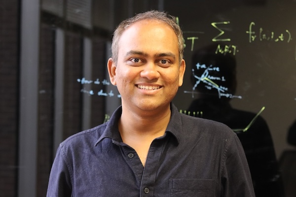 A portrait of Pratik Chaudhari, wearing a dark polo shirt and with a dark glass board with mathematical notation behind him.