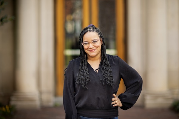 A portrait of Ro Encarnacion, wearing a black long-sleeve top and with one hand on her hip. Her hair is in braids and she has glasses.