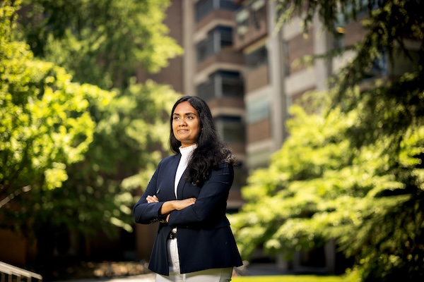 Lasya Sreepada stands with her arms crossed, looking into the distance, with trees and campus buildings in the background