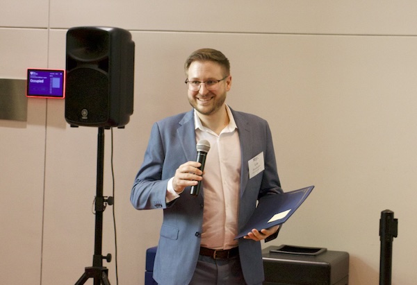 Lyle Brunhofer, wearing a slate suit, holds a microphone while MC'ing Senior Design