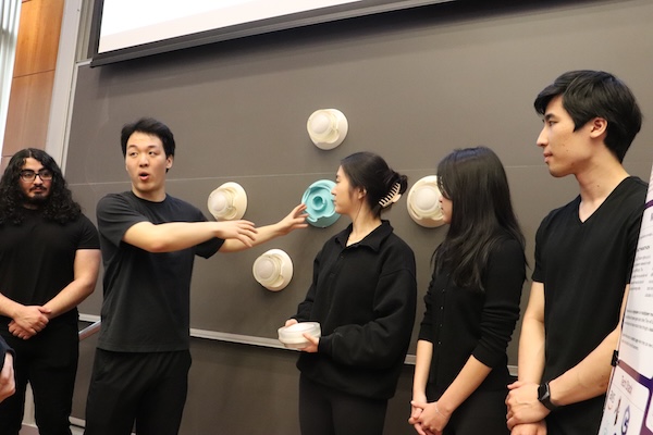 Five students in black tshirts stand before a blackboard. one of them demonstrates while talking.
