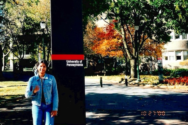 A young woman wearing a denim jacket and jeans stands in front of a sign on UPenn's campus, with fall foliage in the backhround.