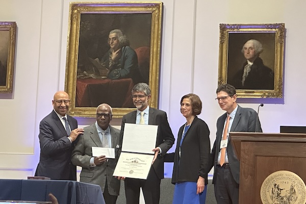 Awardee Vijay Kumar stands flanked by members of the award committee, underneath a portrait of Ben Franklin.