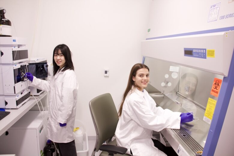 Two women in white lab coats manipulate scientific equipment.