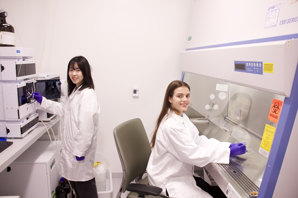 Two women in white lab coats manipulate scientific equipment.