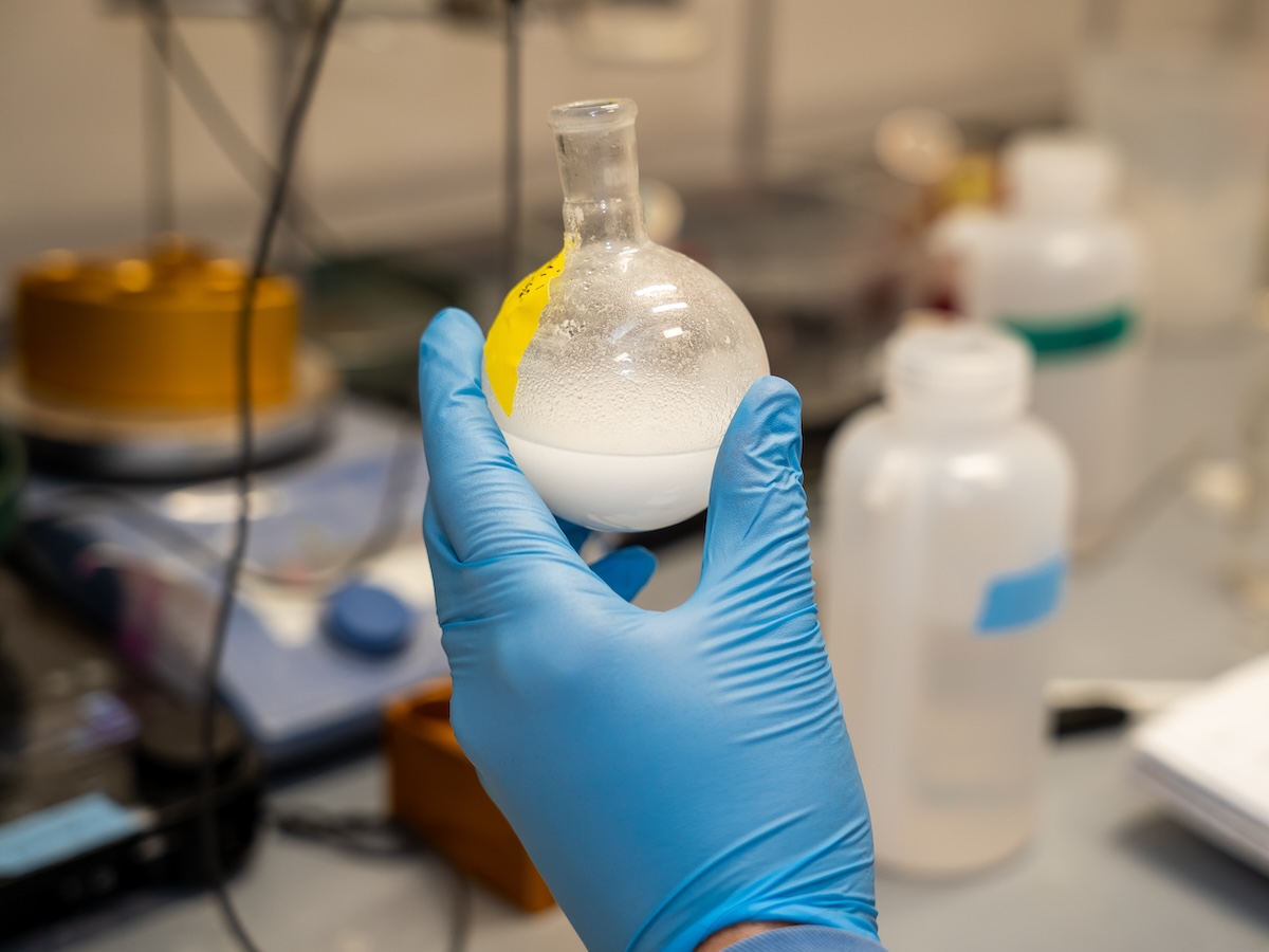 A hand in a blue rubber glove holds a spherical glass container filled with white liquid. 