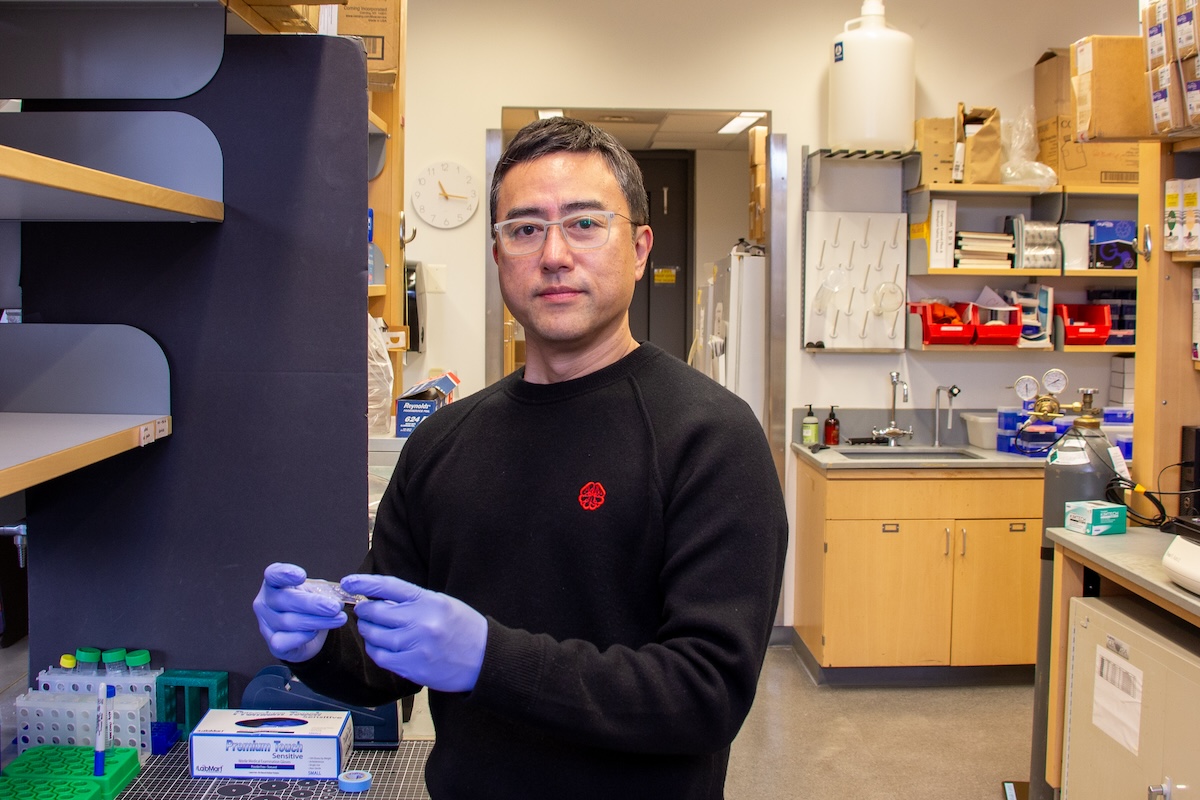 A male scientist in a black crewneck sweater, purple rubber gloves and goggles looks toward the camera while holding a small plastic plate