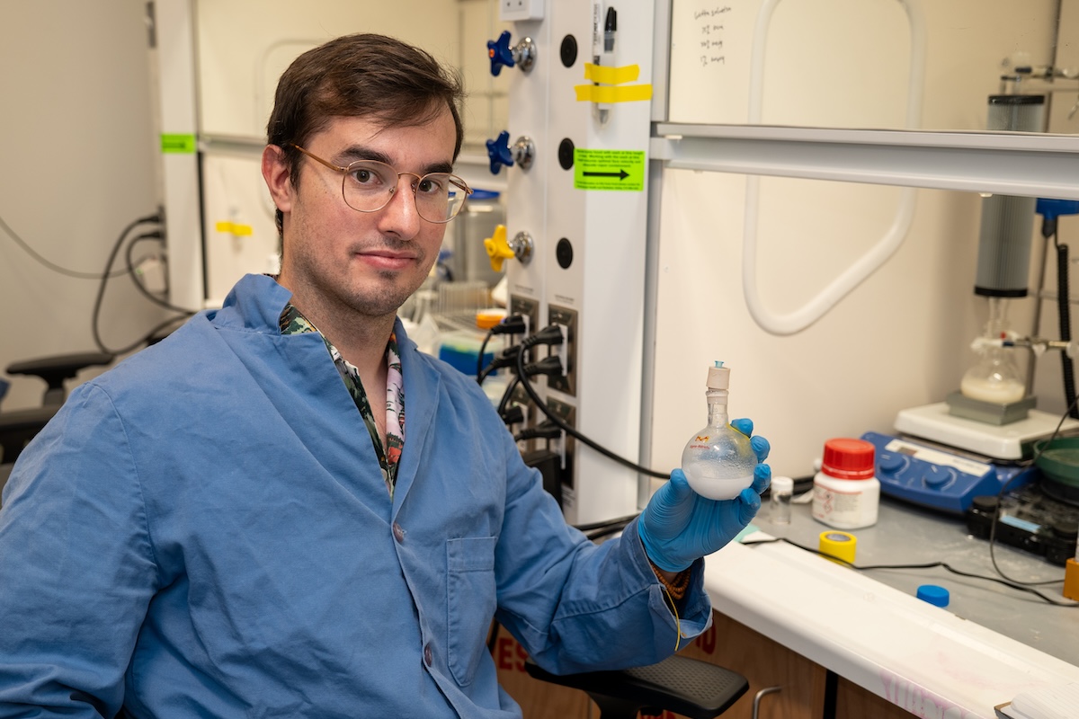 A man in a blue lab coat wearing rubber gloves and glasses holds a spherical container of white liquid and looks towards the camera while sitting in front of a fume hood.