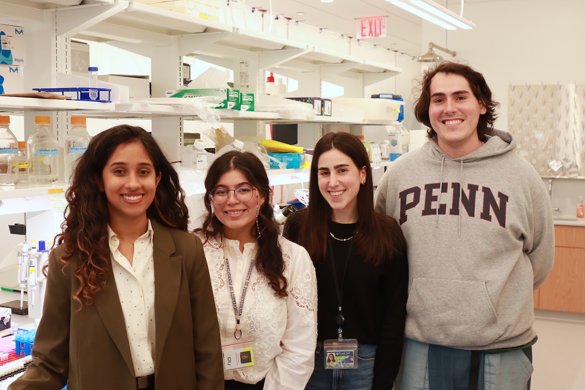 Four researchers, three female and one male, stand in a row smiling in front of their lab bench.