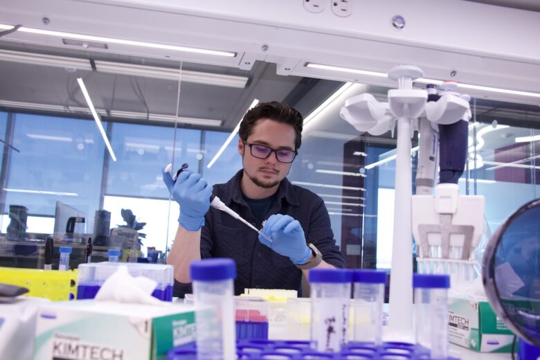 A man uses a micropipette at a lab bench to transfer fluid to a test tube.