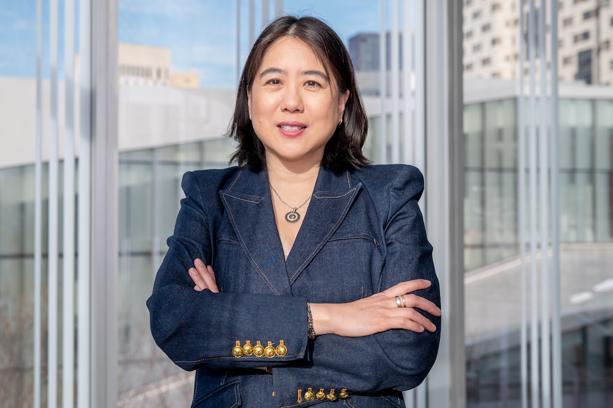 Vanessa Chan stands with her arms crossed in front of a window, wearing a blazer with gold buttons. 