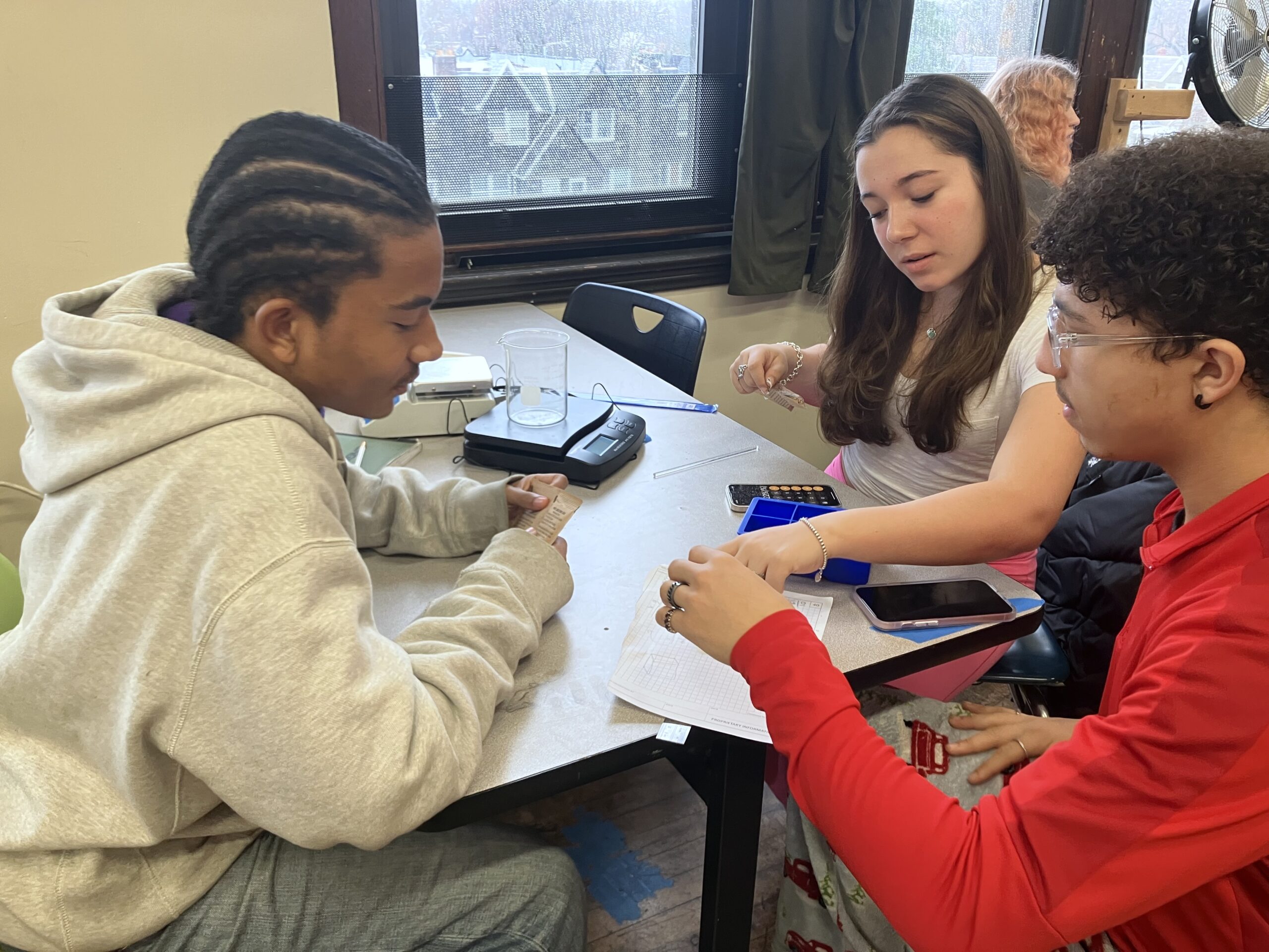 Three teenagers sit at a table with a beaker, scale and calculator, conducting a scientific experiment.