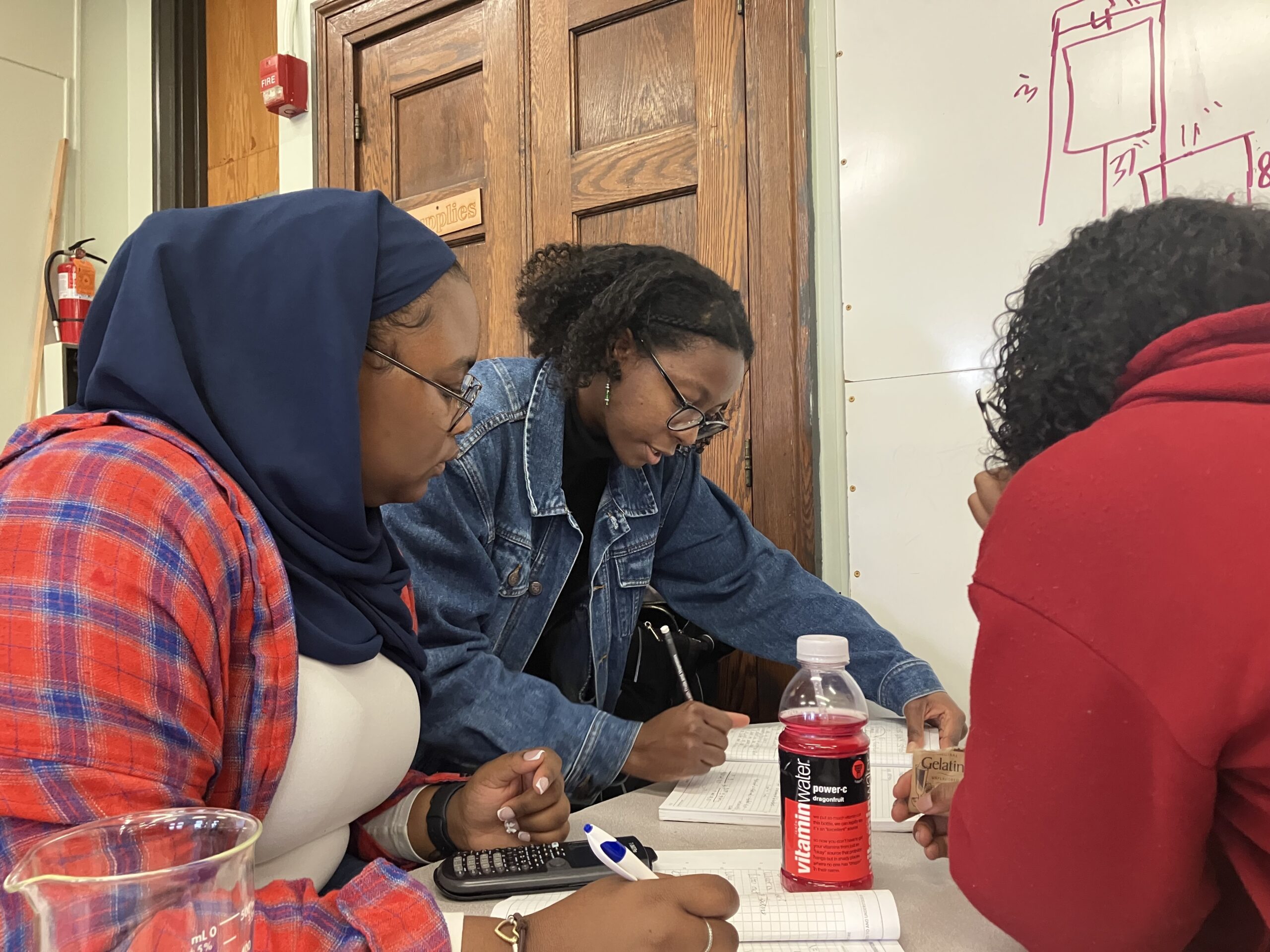 A teacher in a jean jacket writes in a notebook, showing two students how to make a calculation.