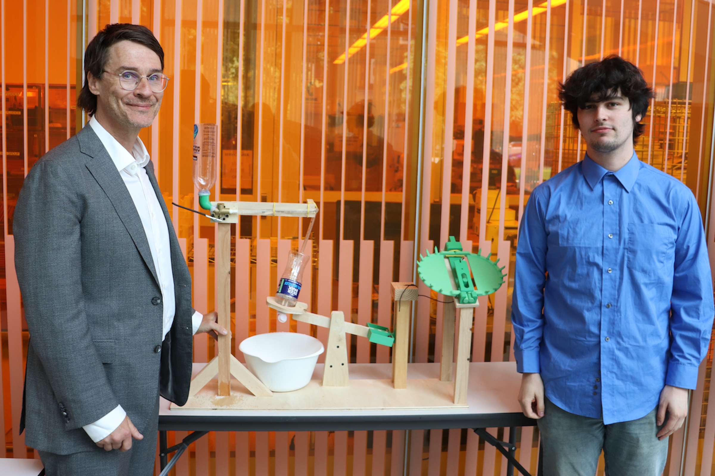 Two men in collared shirts face the camera, flanking a table supporting a wooden model of a Venus fly trap.