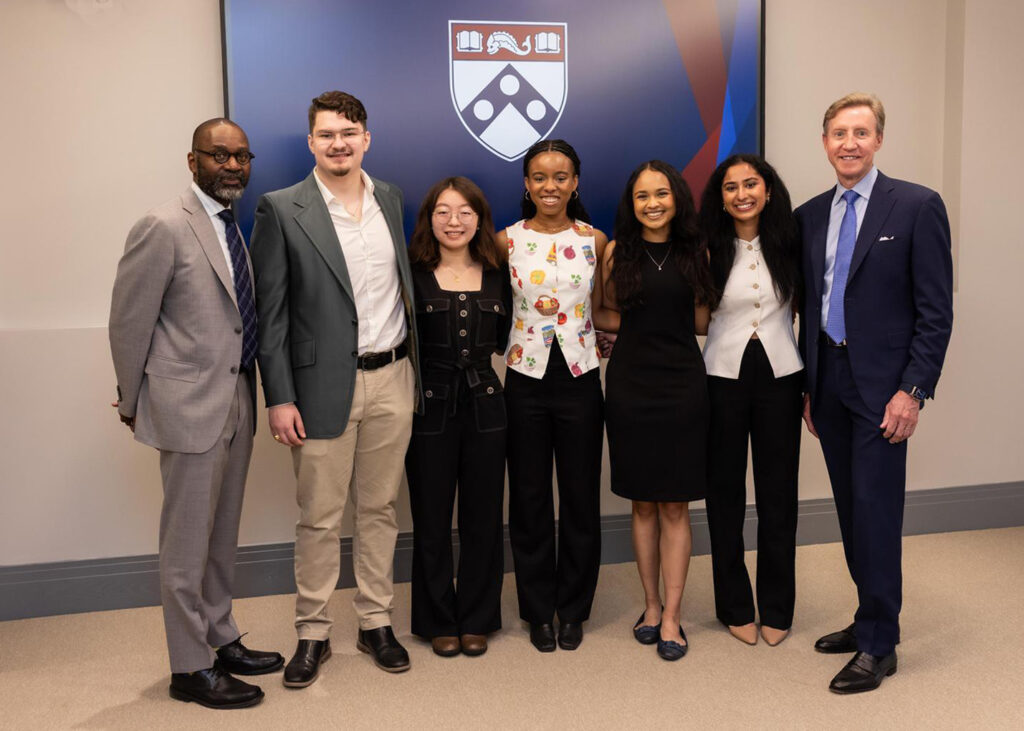 A picture of student prize winners in front of a screen with the Penn logo. From left to right, the people are as follows:(From left) Provost John L. Jackson Jr, Jack Roney, Ejun Mary Hong, Imani Nkrumah Ardayfio, Inaya Zaman, Rashmi Acharya, and Penn President J. Larry Jameson.