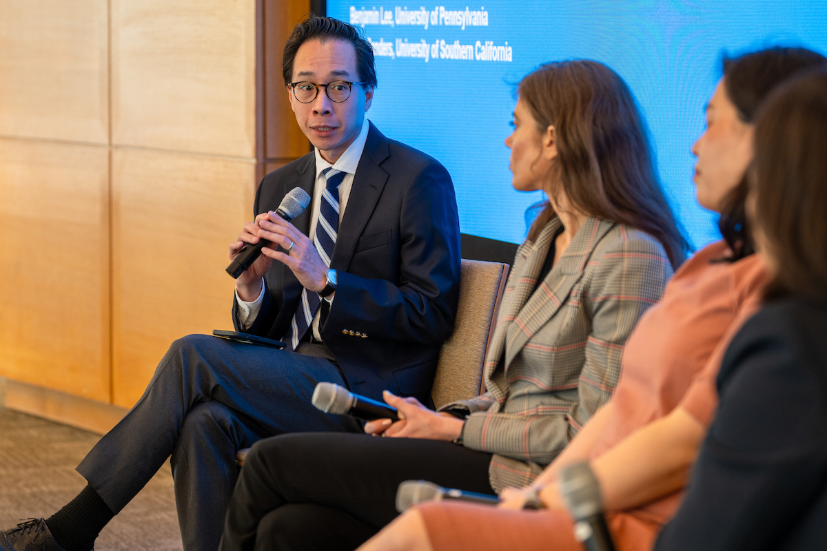 Two women and one man in professional attire sit on a panel on stage and speak using microphones.