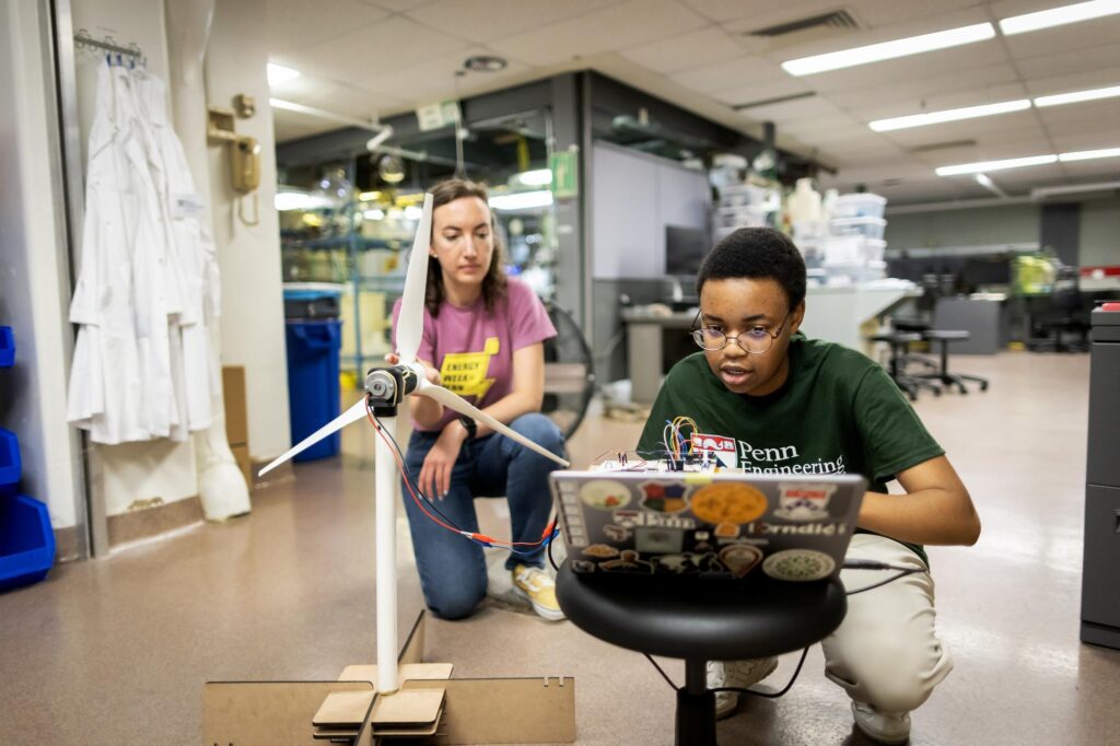 A picture of a professor and student kneeling in a lab in front of a computer and small windmill.