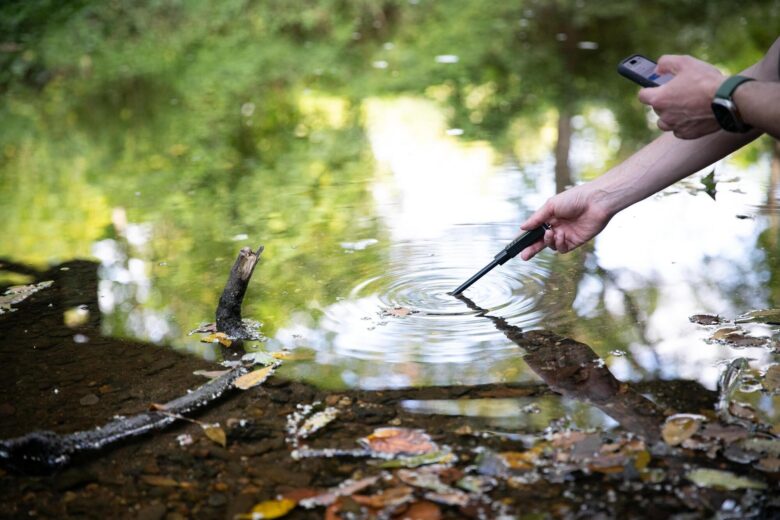 A picture of a creek with someone putting in a test tube to get a sample of the water.