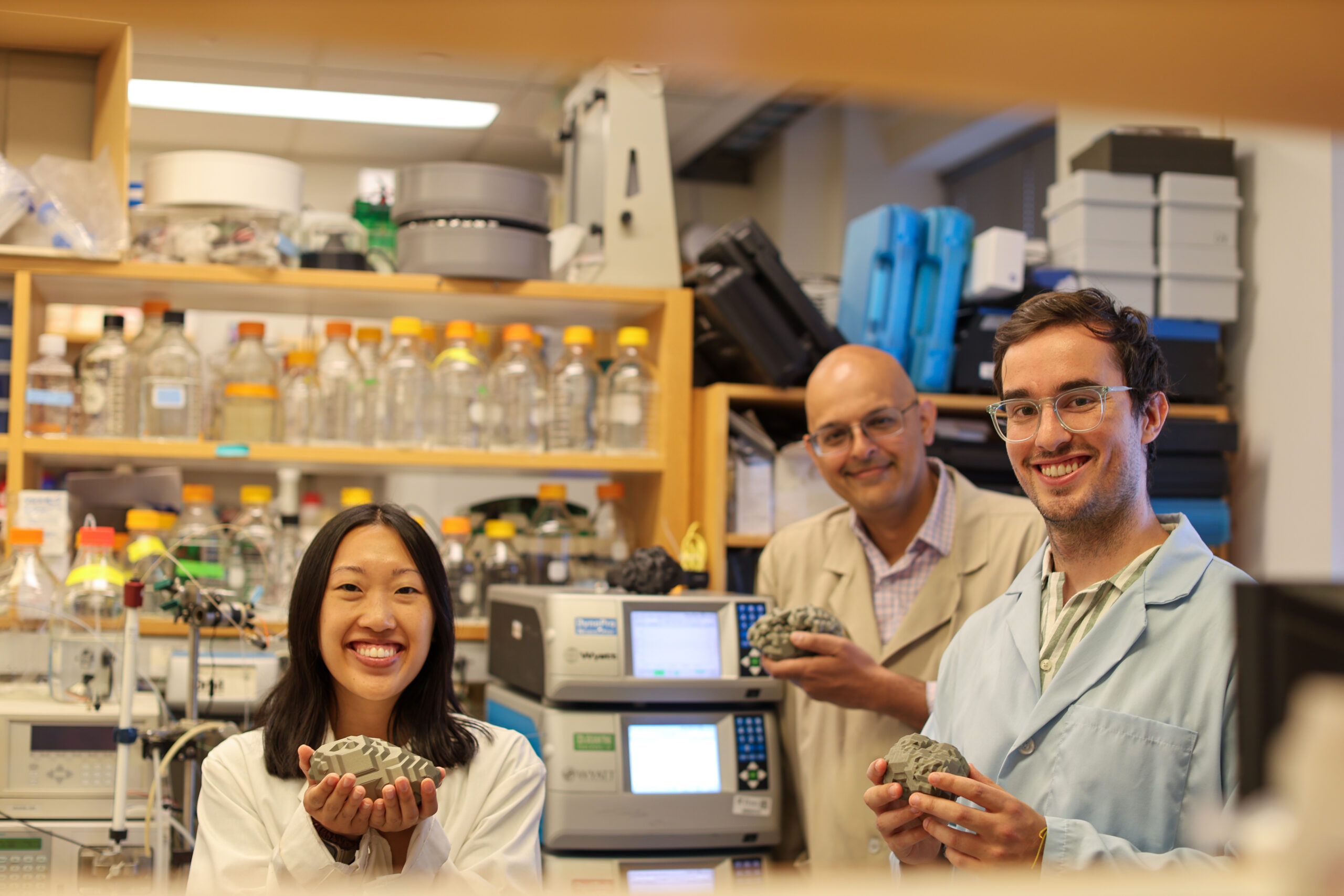 A trio of researchers pose in a lab with 3D printed plastic models of nanoparticles.