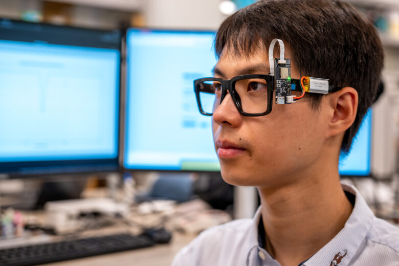 A young man wears a pair of glasses with a device attached to one arm of the glasses.