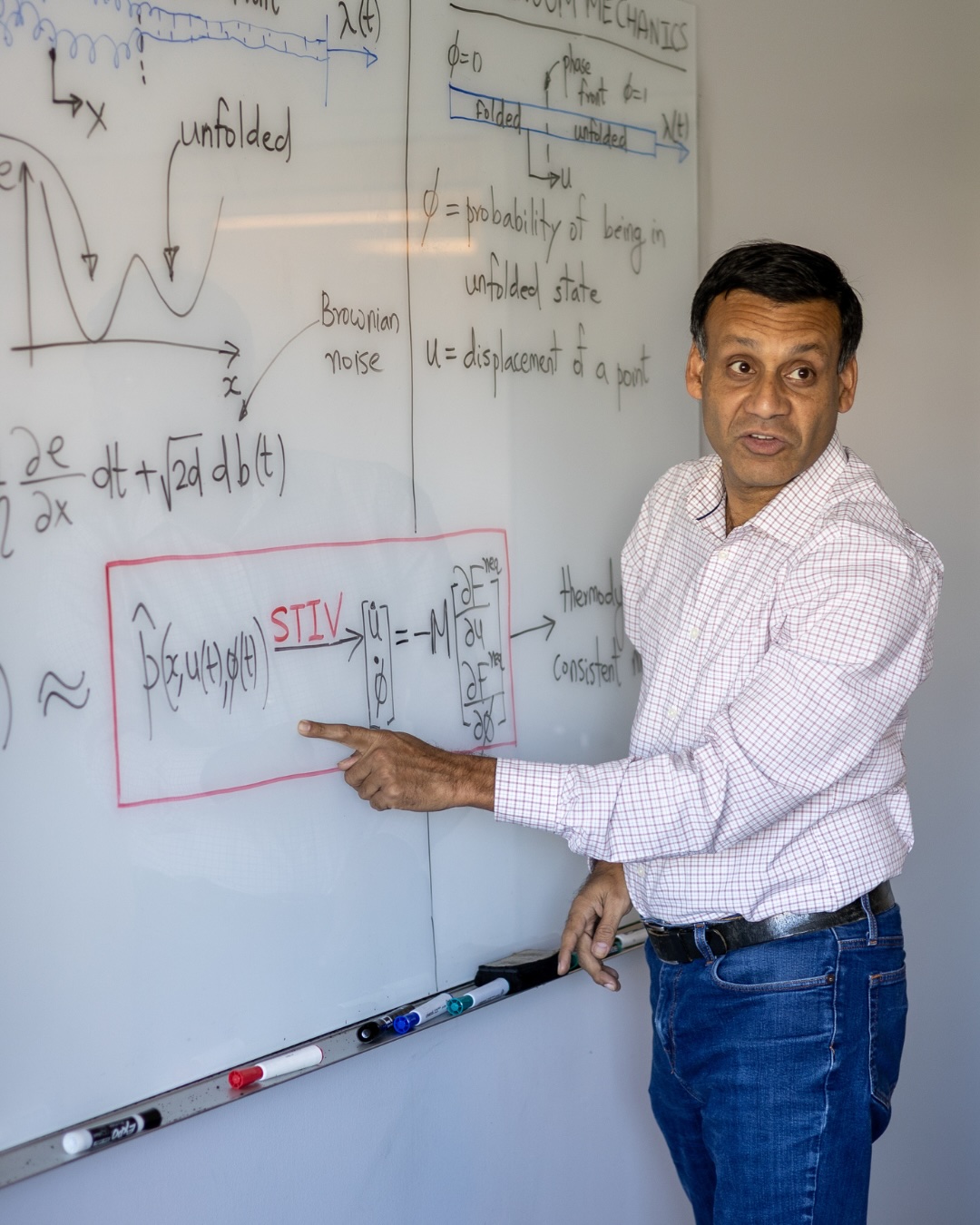 A professor stands in front of a whiteboard covered in math and gestures at it. 